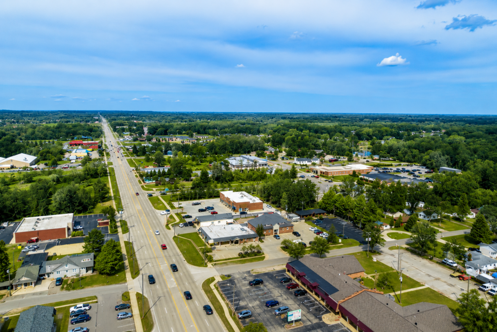 Full HD aerial view of a suburban commercial area with a main road, businesses, parking lots, homes, and green trees under a clear blue sky.